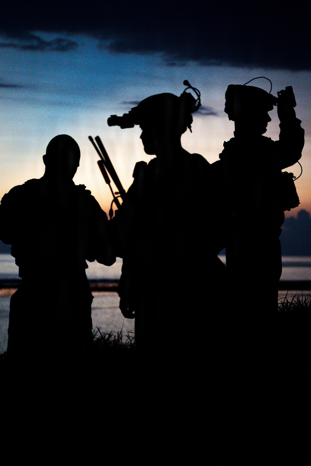 A silhouetted image of three heavily equipped military or tactical personnel photographed against a twilight/sunset sky near a body of water. The figures are rendered entirely in dark silhouette, revealing only their outlines and equipment profiles. The dramatic backlighting creates a high-contrast composition typical of military photography.