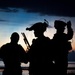 A silhouetted image of three heavily equipped military or tactical personnel photographed against a twilight/sunset sky near a body of water. The figures are rendered entirely in dark silhouette, revealing only their outlines and equipment profiles. The dramatic backlighting creates a high-contrast composition typical of military photography.