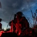 A nighttime scene illuminated by red light showing what appear to be military or tactical personnel crouching in tall grass or vegetation. The image is taken from a low ground-level angle, with a dramatic twilight sky featuring clouds and faint stars visible in the background. The red light source casts strong shadows and silhouettes, creating a high-contrast, atmospheric composition.