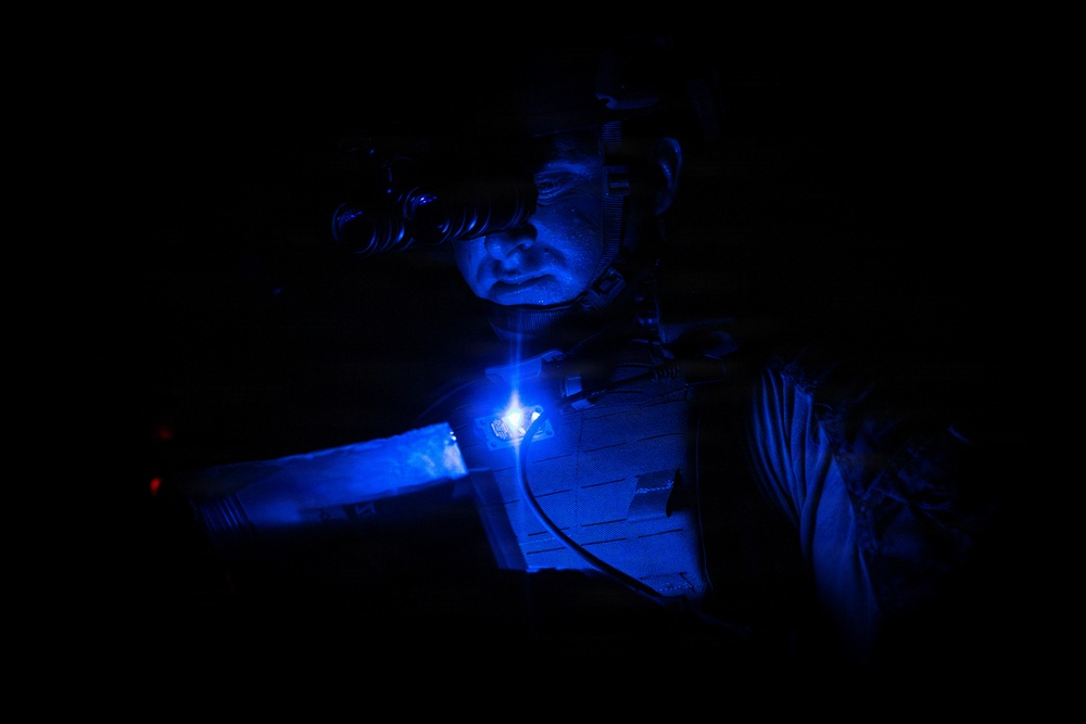 A person wearing night vision goggles and tactical body armor is illuminated by a bright blue light source attached to their chest in an otherwise completely dark environment. The blue light casts a strong glow across their face and equipment, suggesting a nighttime military or law enforcement operation. A small red light is also faintly visible to the left side of the frame.