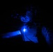 A person wearing night vision goggles and tactical body armor is illuminated by a bright blue light source attached to their chest in an otherwise completely dark environment. The blue light casts a strong glow across their face and equipment, suggesting a nighttime military or law enforcement operation. A small red light is also faintly visible to the left side of the frame.