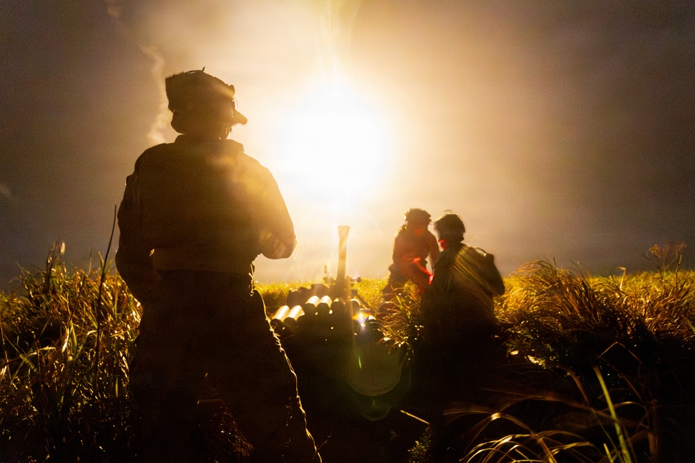 A nighttime military field operation showing silhouetted soldiers in a grassy area illuminated by a bright muzzle flash or explosion in the background. At least three personnel are visible, with what appears to be a mortar tube and stacked ammunition rounds in the foreground. The intense backlighting creates strong silhouettes and lens flare effects.