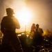 A nighttime military field operation showing silhouetted soldiers in a grassy area illuminated by a bright muzzle flash or explosion in the background. At least three personnel are visible, with what appears to be a mortar tube and stacked ammunition rounds in the foreground. The intense backlighting creates strong silhouettes and lens flare effects.