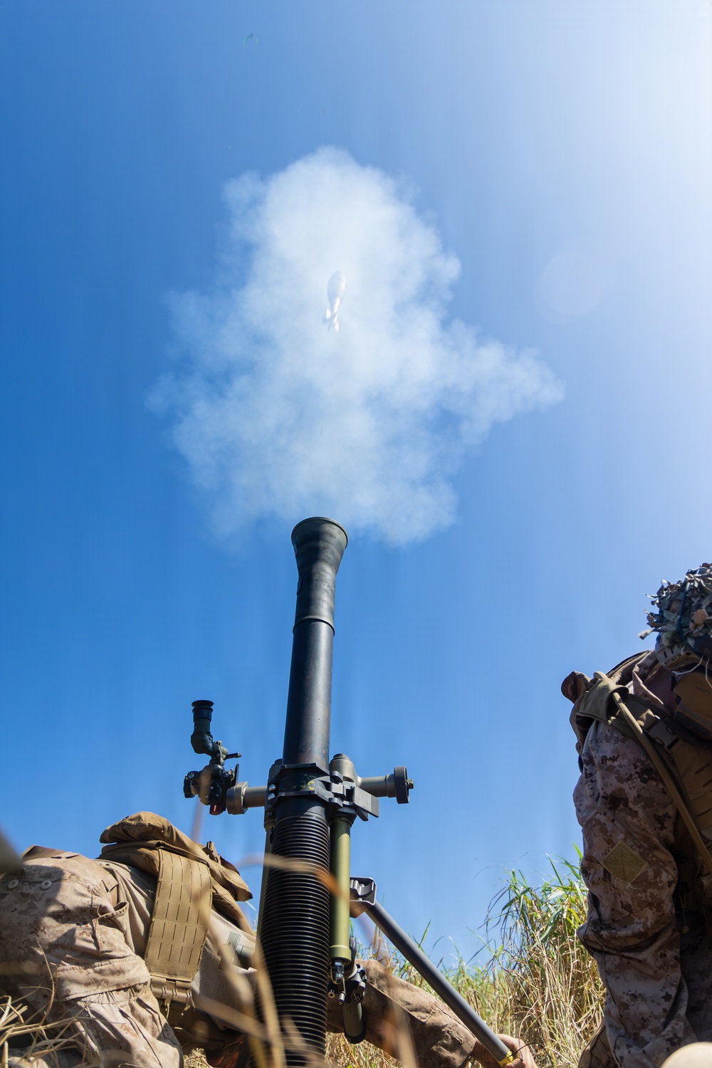 A mortar system has just been fired, with a projectile visibly in flight above the tube against a clear blue sky. White propellant smoke billows from the muzzle, capturing the moment immediately after discharge. At least one soldier in digital camouflage (MARPAT pattern) is visible to the right of the weapon system.