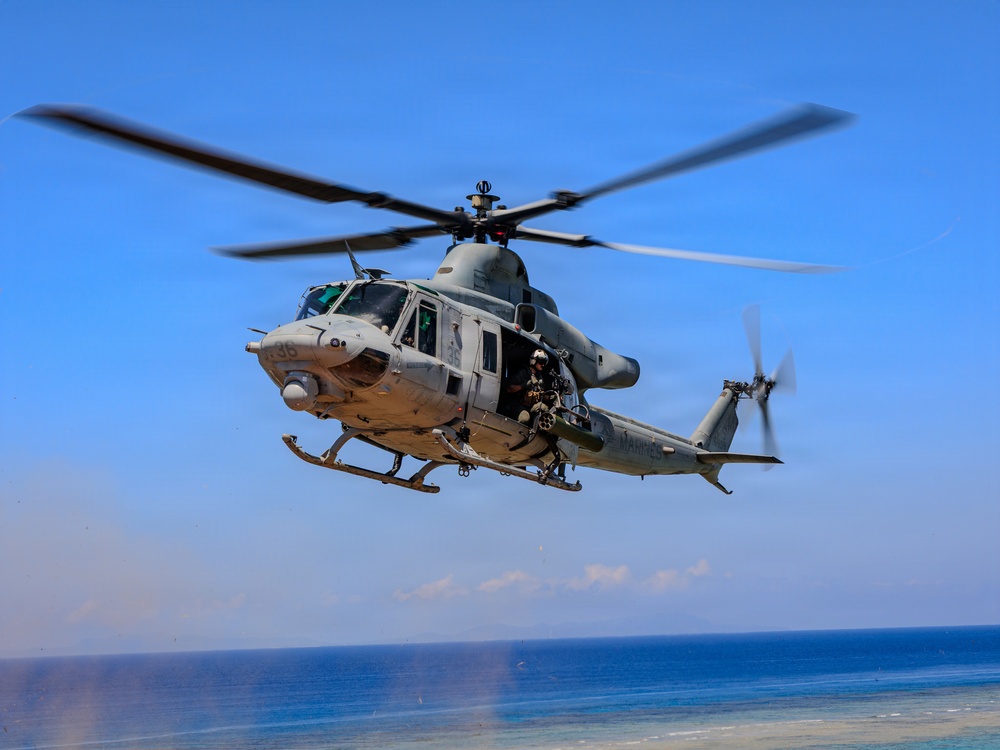 A U.S. Marines military helicopter, identified as a Bell UH-1Y Venom, is captured in flight over a coastal area with blue ocean and sky in the background. The helicopter bears the 'MARINES' marking on its tail boom and the number '36' on its nose. A crew member is visible seated in the open side door, and the main rotor blades show motion blur indicating active flight.