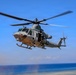 A U.S. Marines military helicopter, identified as a Bell UH-1Y Venom, is captured in flight over a coastal area with blue ocean and sky in the background. The helicopter bears the 'MARINES' marking on its tail boom and the number '36' on its nose. A crew member is visible seated in the open side door, and the main rotor blades show motion blur indicating active flight.