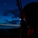 A person is writing in a notebook or document outdoors at night, illuminated by a red light source attached near their head. A rifle or long firearm is visible slung over their shoulder or back. The scene is set against a twilight sky with visible clouds and a moon in the upper left corner.