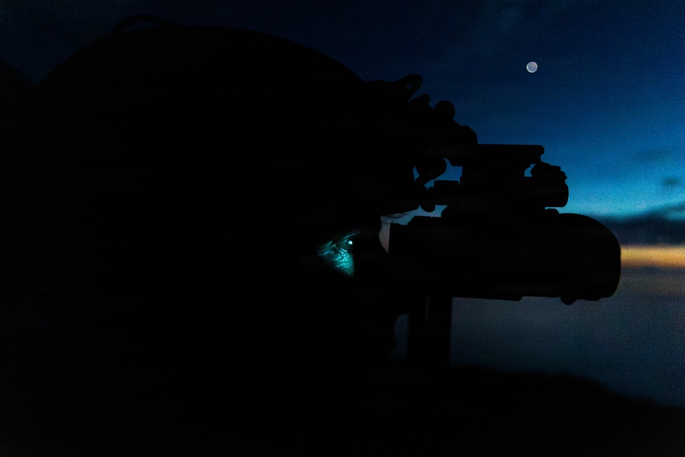 A silhouetted figure is using a rifle with an optical scope during twilight or pre-dawn hours. The person appears to be wearing a helmet equipped with night vision goggles (NVGs), with a faint cyan/teal glow visible at the eye interface. The moon is visible in the upper right portion of the darkening blue sky.