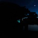 A silhouetted figure is using a rifle with an optical scope during twilight or pre-dawn hours. The person appears to be wearing a helmet equipped with night vision goggles (NVGs), with a faint cyan/teal glow visible at the eye interface. The moon is visible in the upper right portion of the darkening blue sky.