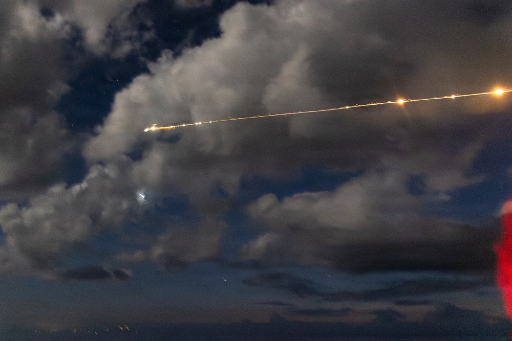 A long-exposure night photograph captures a bright linear trail of lights streaking diagonally across a cloudy sky, consistent with an aircraft or rocket trajectory captured over multiple seconds. The trail shows multiple distinct bright points along its length, suggesting either multiple light sources or stroboscopic capture of a single moving object. A separate glowing object is visible below and to the left of the main trail, possibly a second aircraft or celestial body.