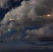 A long-exposure night photograph captures a bright linear trail of lights streaking diagonally across a cloudy sky, consistent with an aircraft or rocket trajectory captured over multiple seconds. The trail shows multiple distinct bright points along its length, suggesting either multiple light sources or stroboscopic capture of a single moving object. A separate glowing object is visible below and to the left of the main trail, possibly a second aircraft or celestial body.
