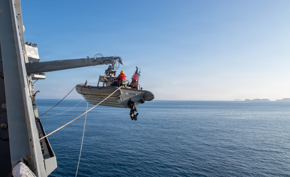 A rigid-hull inflatable boat (RHIB) is being deployed or recovered via a davit crane from the side of a naval vessel over open ocean. At least two personnel are visible aboard the RHIB, one wearing a red jacket and yellow helmet, and an American flag is mounted on the boat. A person in dark tactical gear is suspended below the RHIB, hanging from a line.