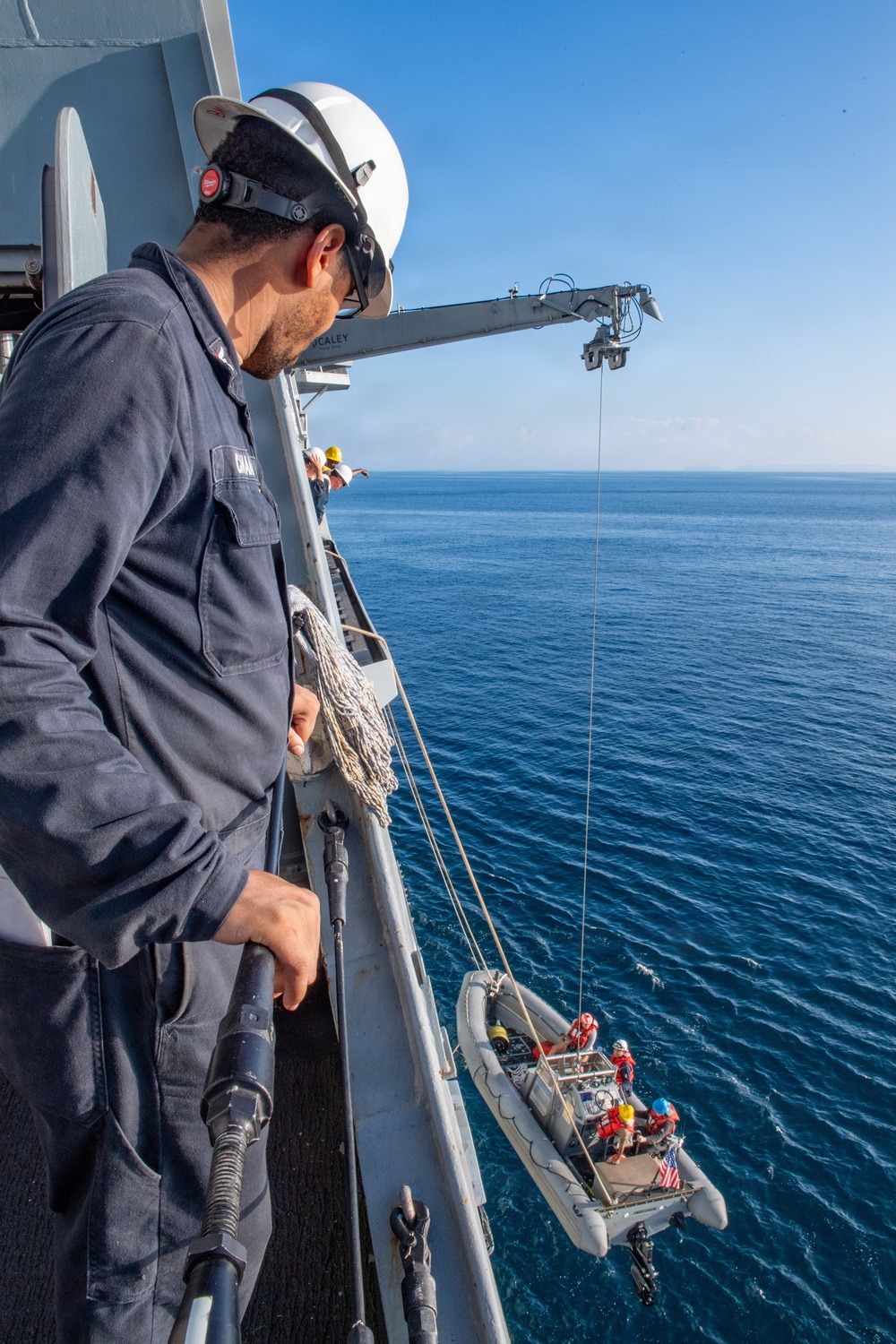A crew member wearing a white hard hat and hearing protection stands on the deck of a vessel, overseeing the lowering of a rigid inflatable boat (RIB) via a davit crane over open ocean. The RIB contains several personnel in safety gear and displays a small American flag. The crane arm is partially labeled, appearing to read 'HEALEY', suggesting this may be the USCGC Healy or a similarly named vessel.