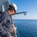 A crew member wearing a white hard hat and hearing protection stands on the deck of a vessel, overseeing the lowering of a rigid inflatable boat (RIB) via a davit crane over open ocean. The RIB contains several personnel in safety gear and displays a small American flag. The crane arm is partially labeled, appearing to read 'HEALEY', suggesting this may be the USCGC Healy or a similarly named vessel.