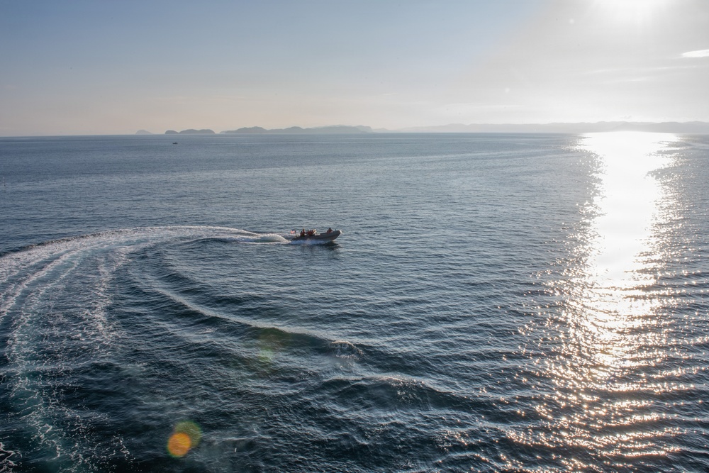 An aerial or elevated view of an open ocean scene featuring a rigid inflatable boat (RIB) making a curved turn, leaving a visible white wake trail on the water's surface. The sun is low on the horizon to the right, creating a bright glare path across the water. Distant islands or landmasses are faintly visible along the horizon in the background.