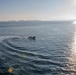 An aerial or elevated view of an open ocean scene featuring a rigid inflatable boat (RIB) making a curved turn, leaving a visible white wake trail on the water's surface. The sun is low on the horizon to the right, creating a bright glare path across the water. Distant islands or landmasses are faintly visible along the horizon in the background.