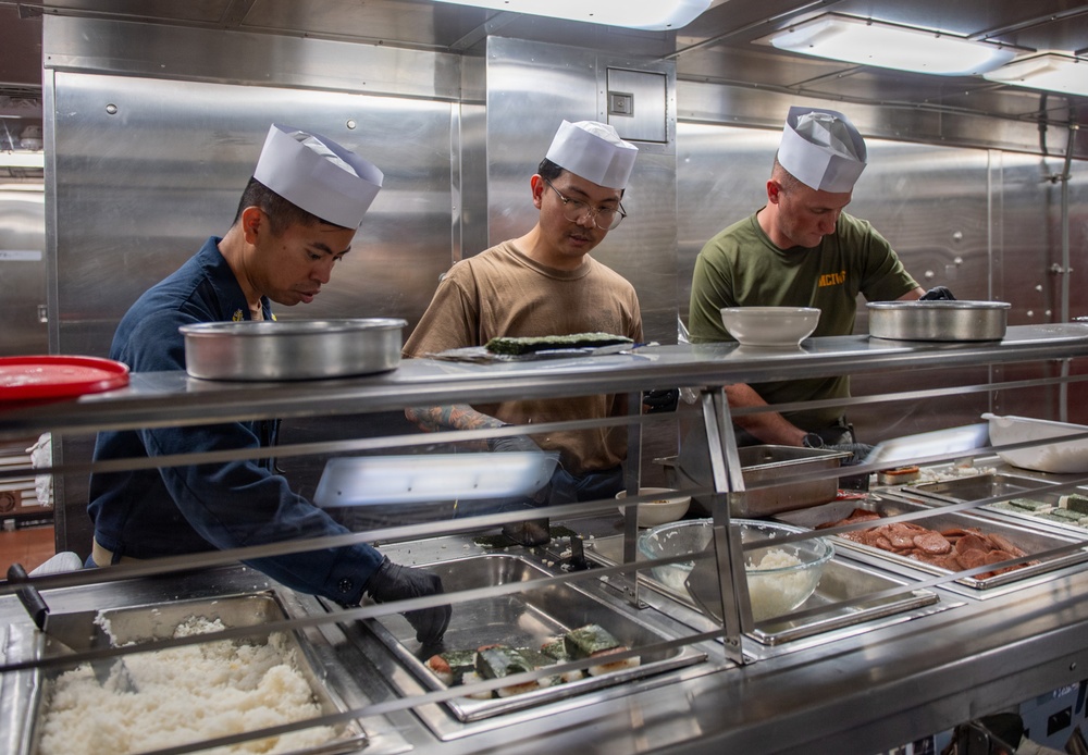 Three individuals wearing white paper chef hats are working behind a food service counter in what appears to be a military galley or mess hall. They are preparing and serving food items including rice, sushi rolls, and other dishes arranged in stainless steel serving trays. The workers are dressed in different military-style uniforms including navy blue, tan, and olive green shirts.