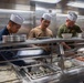 Three individuals wearing white paper chef hats are working behind a food service counter in what appears to be a military galley or mess hall. They are preparing and serving food items including rice, sushi rolls, and other dishes arranged in stainless steel serving trays. The workers are dressed in different military-style uniforms including navy blue, tan, and olive green shirts.