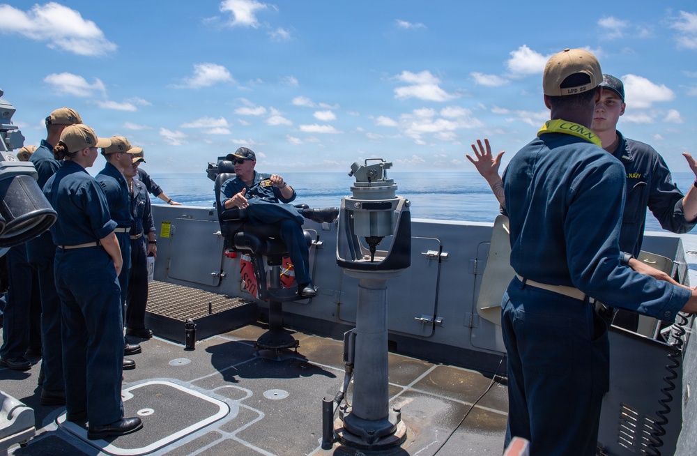 A group of U.S. Navy personnel are gathered on the open deck of a naval vessel, appearing to receive instruction or a briefing from a senior sailor seated in a command chair. The scene takes place at sea under clear blue skies with calm ocean waters visible in the background. Personnel are wearing standard Navy working uniforms with tan caps, and one individual has a yellow 'CONN' lanyard visible.