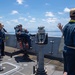 A group of U.S. Navy personnel are gathered on the open deck of a naval vessel, appearing to receive instruction or a briefing from a senior sailor seated in a command chair. The scene takes place at sea under clear blue skies with calm ocean waters visible in the background. Personnel are wearing standard Navy working uniforms with tan caps, and one individual has a yellow 'CONN' lanyard visible.