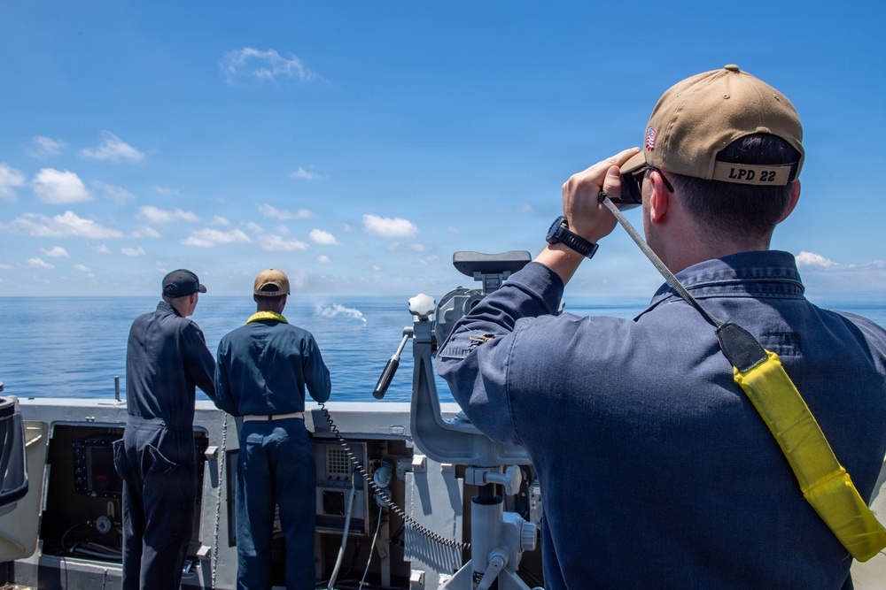 Three naval personnel are stationed on the deck of a U.S. Navy ship, designated LPD 22, observing the open ocean. The foreground sailor is using binoculars or a rangefinder device and wears a cap marked 'LPD 22' with a yellow safety lanyard. Two other sailors stand at the ship's railing looking toward what appears to be a small splash or disturbance on the water's surface.