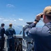 Three naval personnel are stationed on the deck of a U.S. Navy ship, designated LPD 22, observing the open ocean. The foreground sailor is using binoculars or a rangefinder device and wears a cap marked 'LPD 22' with a yellow safety lanyard. Two other sailors stand at the ship's railing looking toward what appears to be a small splash or disturbance on the water's surface.