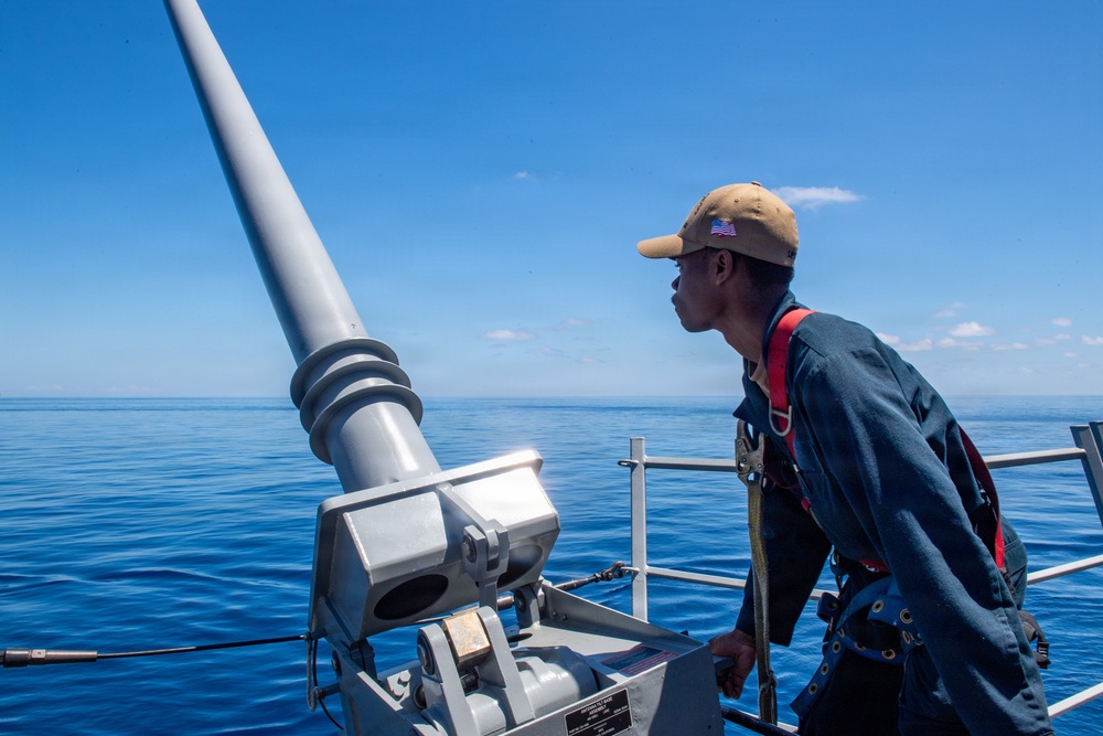 A naval service member stands on the deck of a military vessel, looking out over calm, deep blue ocean waters. The individual is positioned near what appears to be a deck-mounted gun or weapon system, wearing a tan cap with a visible American flag patch and a blue coverall uniform with a safety harness. The scene is set under clear, bright blue skies with minimal cloud cover.