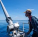 A naval service member stands on the deck of a military vessel, looking out over calm, deep blue ocean waters. The individual is positioned near what appears to be a deck-mounted gun or weapon system, wearing a tan cap with a visible American flag patch and a blue coverall uniform with a safety harness. The scene is set under clear, bright blue skies with minimal cloud cover.