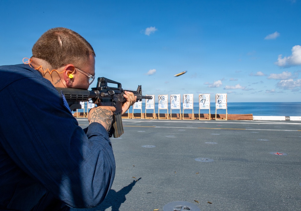 A person in a blue uniform is firing an M16/M4-style rifle on what appears to be the flight deck of a naval vessel, with a spent brass casing visibly ejecting from the weapon. Multiple numbered paper targets are set up downrange against a backdrop of open ocean and blue sky. This appears to be a military marksmanship qualification or training exercise conducted at sea.