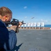 A person in a blue uniform is firing an M16/M4-style rifle on what appears to be the flight deck of a naval vessel, with a spent brass casing visibly ejecting from the weapon. Multiple numbered paper targets are set up downrange against a backdrop of open ocean and blue sky. This appears to be a military marksmanship qualification or training exercise conducted at sea.