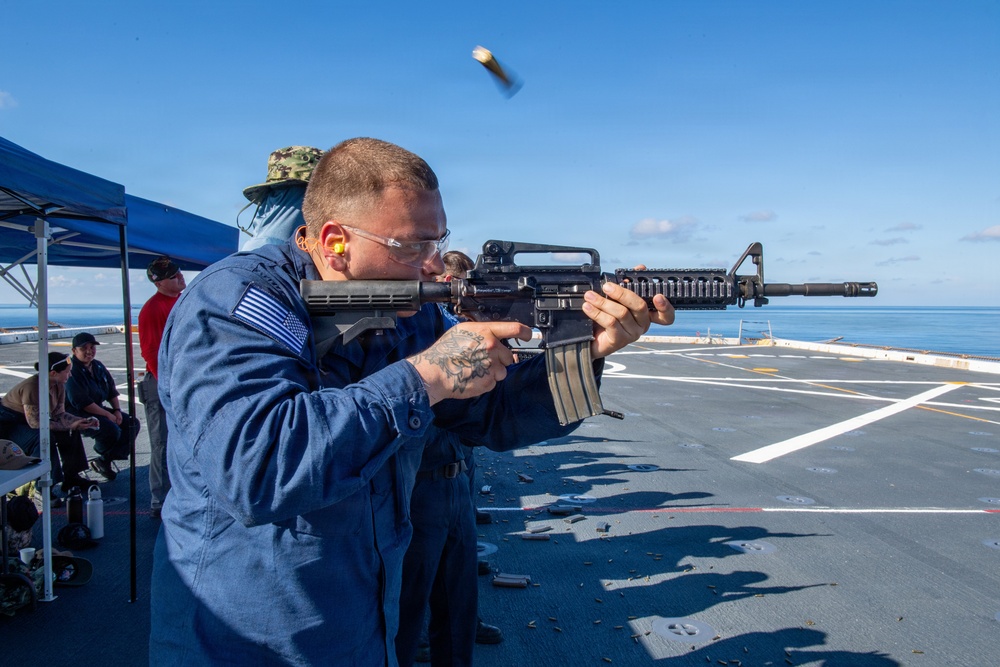 A U.S. Navy sailor in blue coveralls is firing an M16/M4-style rifle on the flight deck of a naval vessel at sea. A spent brass casing is visibly ejecting from the weapon, captured mid-air by the camera. Other personnel are observing from a shaded area under a blue canopy in the background.