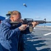 A U.S. Navy sailor in blue coveralls is firing an M16/M4-style rifle on the flight deck of a naval vessel at sea. A spent brass casing is visibly ejecting from the weapon, captured mid-air by the camera. Other personnel are observing from a shaded area under a blue canopy in the background.