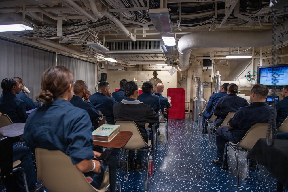 A group of military personnel in navy blue uniforms are seated in a shipboard compartment attending what appears to be a religious service or chapel session. A speaker stands at the front of the room near a red podium, with a screen displaying the word 'SERMON' visible to the right. The setting is clearly aboard a naval vessel, indicated by the exposed pipes, ducts, and industrial ceiling infrastructure.
