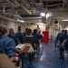 A group of military personnel in navy blue uniforms are seated in a shipboard compartment attending what appears to be a religious service or chapel session. A speaker stands at the front of the room near a red podium, with a screen displaying the word 'SERMON' visible to the right. The setting is clearly aboard a naval vessel, indicated by the exposed pipes, ducts, and industrial ceiling infrastructure.