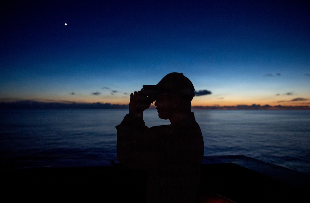 A silhouetted figure wearing a cap is using binoculars or a handheld optical device while standing on what appears to be a vessel at sea during twilight. The scene is backlit by a dramatic sunset or sunrise horizon over calm ocean waters, with a single celestial body visible in the upper sky. The image is a high-contrast silhouette composition emphasizing mood over detail.