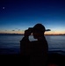 A silhouetted figure wearing a cap is using binoculars or a handheld optical device while standing on what appears to be a vessel at sea during twilight. The scene is backlit by a dramatic sunset or sunrise horizon over calm ocean waters, with a single celestial body visible in the upper sky. The image is a high-contrast silhouette composition emphasizing mood over detail.