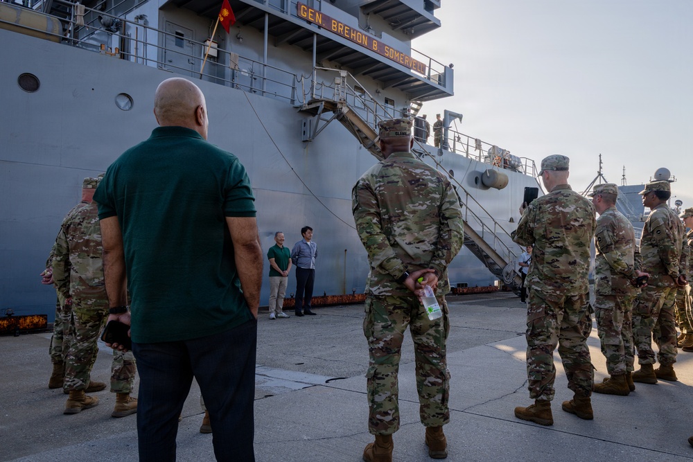 Military personnel and civilians are gathered on a dock in front of a large gray vessel named 'Gen. Brehon B. Somervell.' The group appears to be attending a formal dockside event or briefing near the ship's gangway. Multiple soldiers in OCP (Operational Camouflage Pattern) uniforms are present alongside civilians in casual attire.