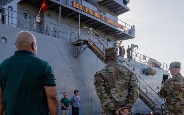 Military personnel and civilians are gathered on a dock in front of a large gray vessel named 'Gen. Brehon B. Somervell.' The group appears to be attending a formal dockside event or briefing near the ship's gangway. Multiple soldiers in OCP (Operational Camouflage Pattern) uniforms are present alongside civilians in casual attire.