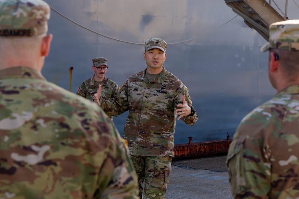 A U.S. Army Colonel (identifiable by the eagle insignia on his chest) is addressing a small group of soldiers in an outdoor industrial setting. He appears to be conducting a briefing or informal discussion, gesturing with both hands while speaking. The setting appears to be near large metal storage tanks or industrial structures.