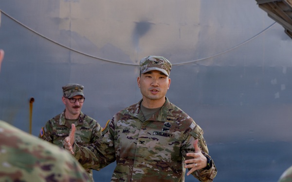 A U.S. Army Colonel (identifiable by the eagle insignia on his chest) is addressing a small group of soldiers in an outdoor industrial setting. He appears to be conducting a briefing or informal discussion, gesturing with both hands while speaking. The setting appears to be near large metal storage tanks or industrial structures.