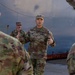 A U.S. Army Colonel (identifiable by the eagle insignia on his chest) is addressing a small group of soldiers in an outdoor industrial setting. He appears to be conducting a briefing or informal discussion, gesturing with both hands while speaking. The setting appears to be near large metal storage tanks or industrial structures.
