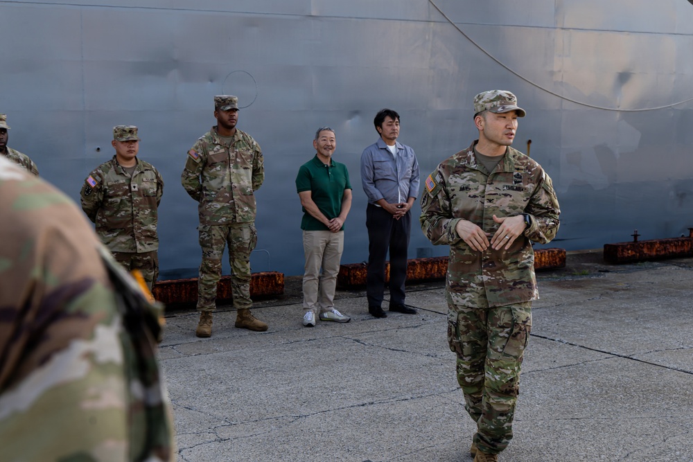 A group of U.S. Army soldiers in OCP (Operational Camouflage Pattern) uniforms are gathered near a large gray vessel or structure at what appears to be a port or dock facility. Two civilians in casual attire are present among the military personnel, suggesting a joint briefing or coordination meeting. A soldier in the foreground appears to be addressing the group.