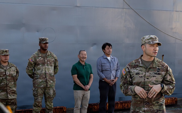 A group of U.S. Army soldiers in OCP (Operational Camouflage Pattern) uniforms are gathered near a large gray vessel or structure at what appears to be a port or dock facility. Two civilians in casual attire are present among the military personnel, suggesting a joint briefing or coordination meeting. A soldier in the foreground appears to be addressing the group.