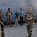 A group of U.S. Army soldiers in OCP (Operational Camouflage Pattern) uniforms are gathered near a large gray vessel or structure at what appears to be a port or dock facility. Two civilians in casual attire are present among the military personnel, suggesting a joint briefing or coordination meeting. A soldier in the foreground appears to be addressing the group.