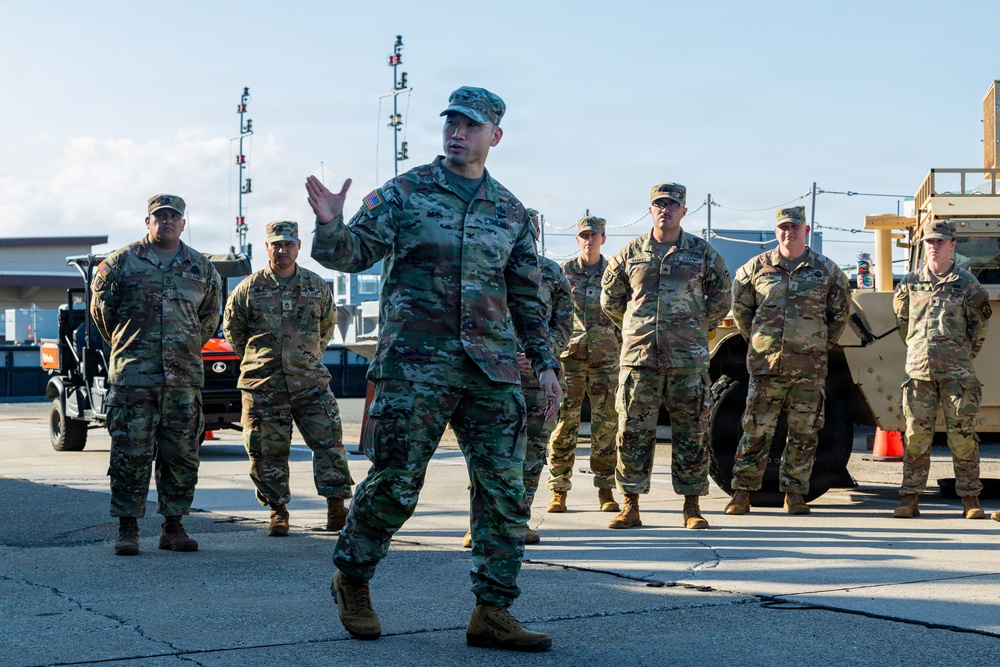 A military briefing or instruction session is taking place outdoors on what appears to be a tarmac or military installation. One soldier in the foreground is actively speaking and gesturing with his hand while a group of approximately 8-10 soldiers stand attentively behind him. Military vehicles and equipment are visible in the background.