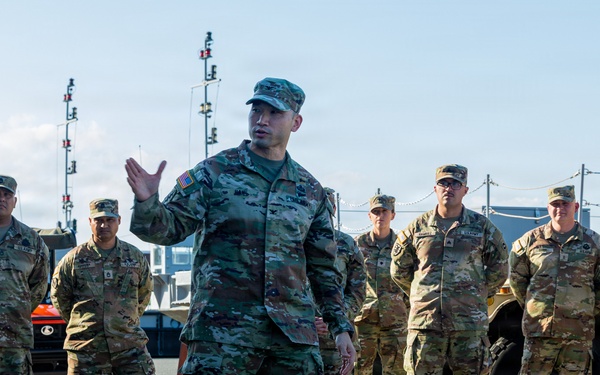 A military briefing or instruction session is taking place outdoors on what appears to be a tarmac or military installation. One soldier in the foreground is actively speaking and gesturing with his hand while a group of approximately 8-10 soldiers stand attentively behind him. Military vehicles and equipment are visible in the background.