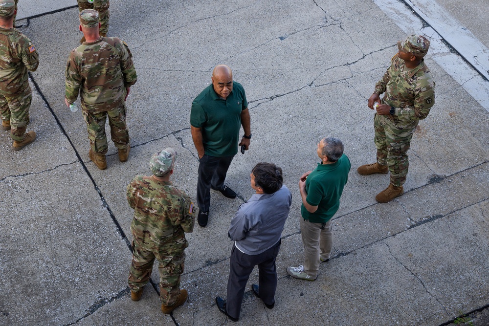An aerial view of a group of individuals gathered on a cracked concrete surface, consisting of military personnel in camouflage uniforms and civilians in casual attire. The group appears to be engaged in a briefing or discussion, with some individuals facing each other in conversation. The photo is taken from an elevated angle, likely from a building or elevated platform.