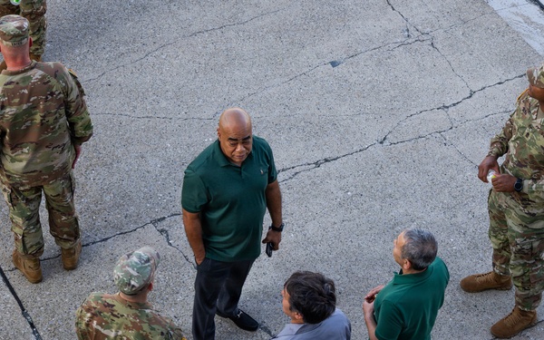 An aerial view of a group of individuals gathered on a cracked concrete surface, consisting of military personnel in camouflage uniforms and civilians in casual attire. The group appears to be engaged in a briefing or discussion, with some individuals facing each other in conversation. The photo is taken from an elevated angle, likely from a building or elevated platform.