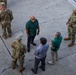 An aerial view of a group of individuals gathered on a cracked concrete surface, consisting of military personnel in camouflage uniforms and civilians in casual attire. The group appears to be engaged in a briefing or discussion, with some individuals facing each other in conversation. The photo is taken from an elevated angle, likely from a building or elevated platform.
