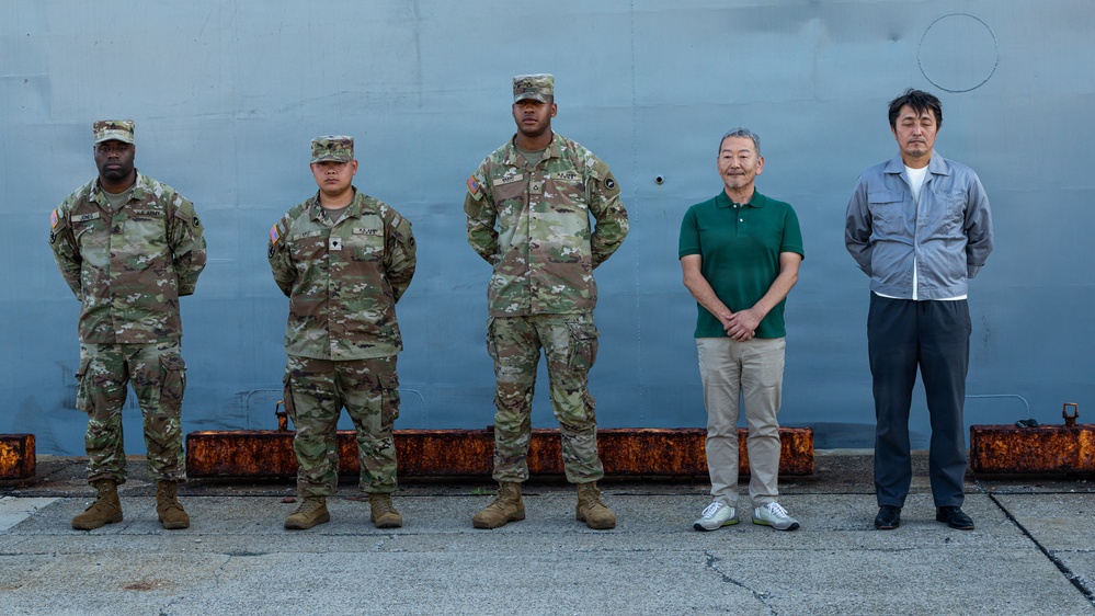 Six individuals stand in a line against a large gray metal structure, likely a ship hull or military vessel at a dock or port facility. Three individuals on the left are wearing U.S. Army OCP (Operational Camouflage Pattern) uniforms with patrol caps, while two individuals on the right are dressed in civilian clothing. The setting appears to be an official or ceremonial group photo opportunity.